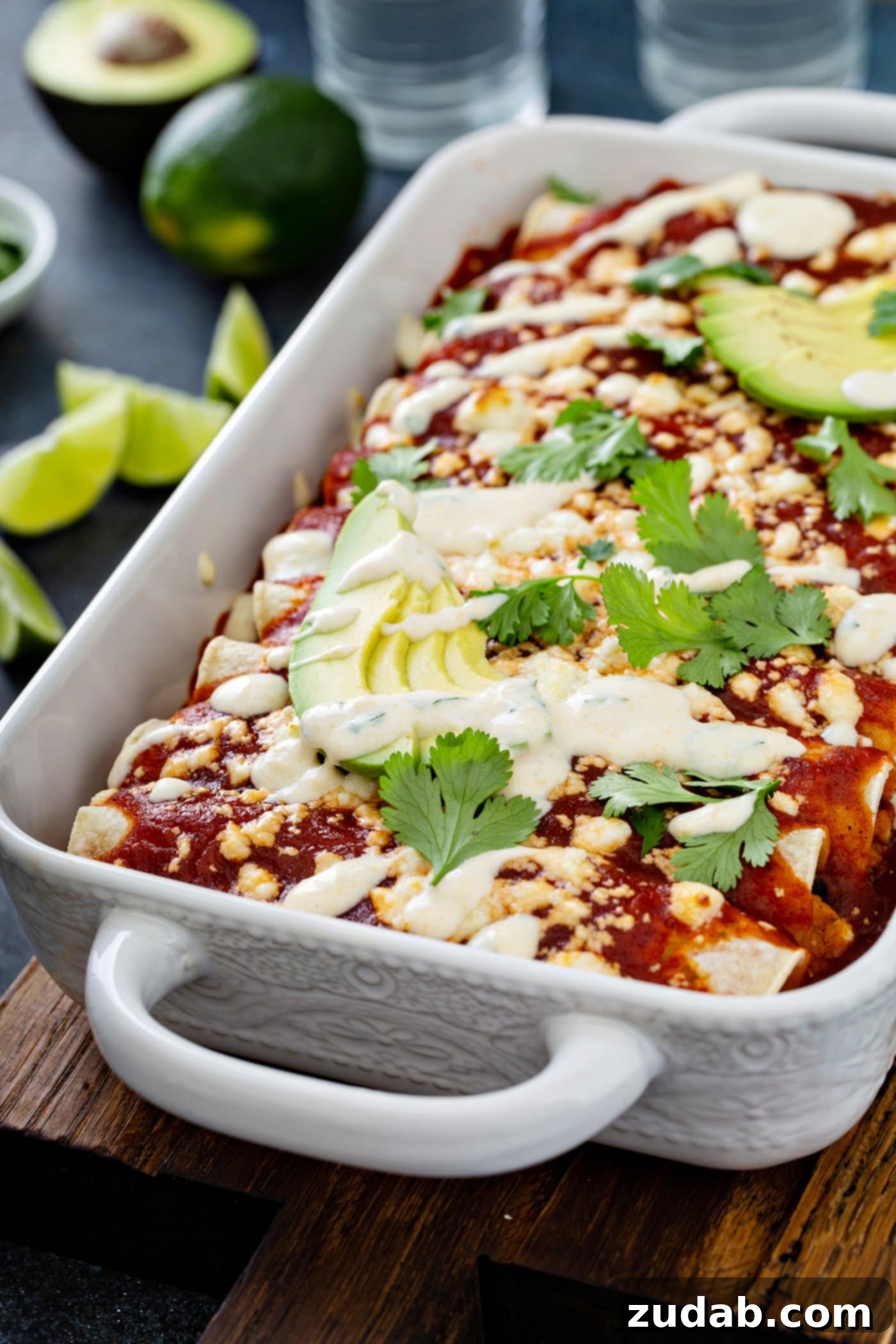 A baking dish with baked sweet potato enchiladas, beautifully garnished. They feature a rich red sauce, crumbled queso fresco cheese, fresh avocado slices, and cilantro leaves, with a delicate drizzle of sweet corn crema over the entire dish.