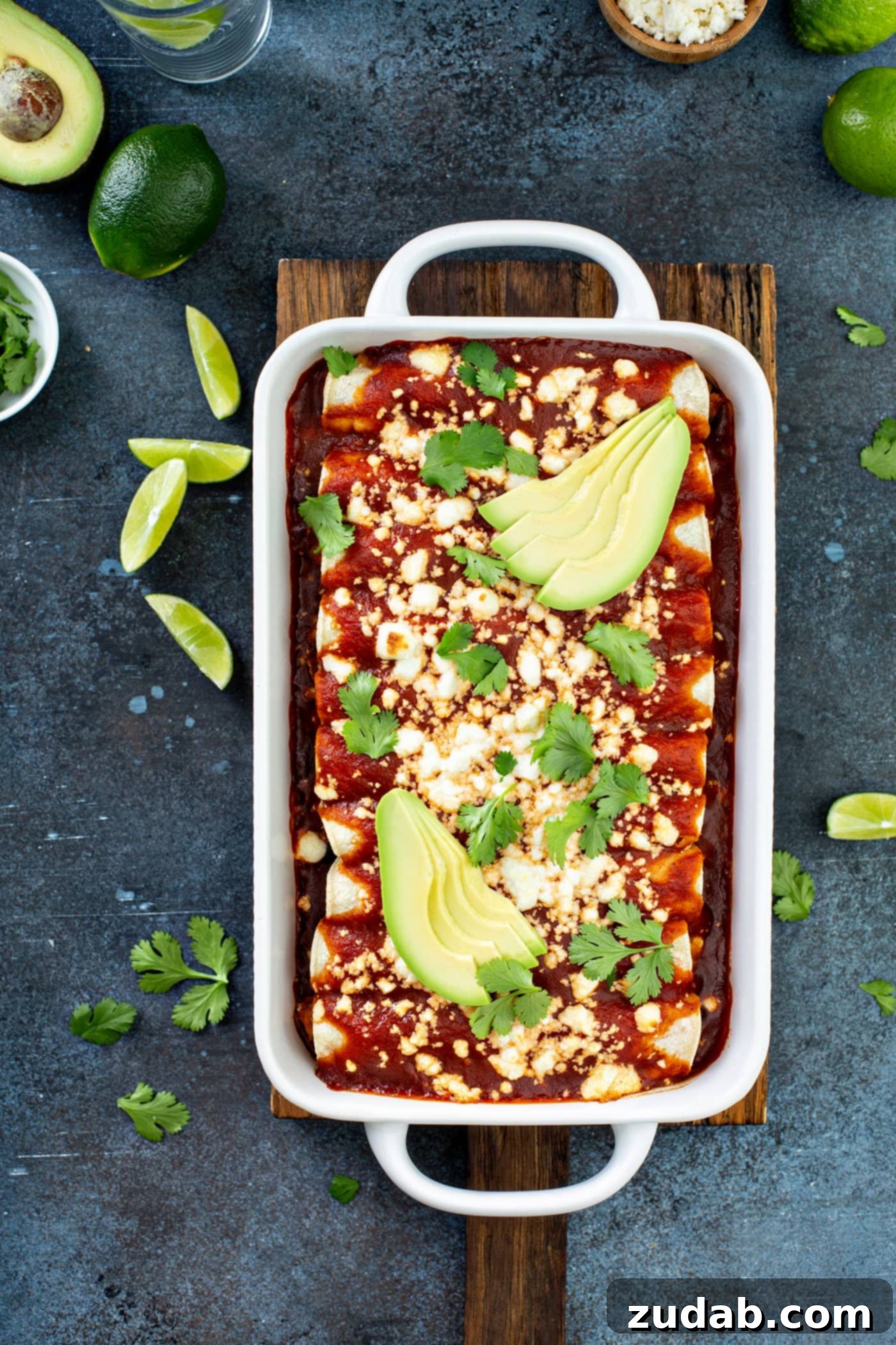 A white casserole dish filled with sweet potato enchiladas. They have red enchilada sauce poured over the top and queso fresco cheese sprinkled on top of the sauce. Then, a few slices of avocado and cilantro leaves top it all off. There are lime wedges and cilantro leaves on the table.