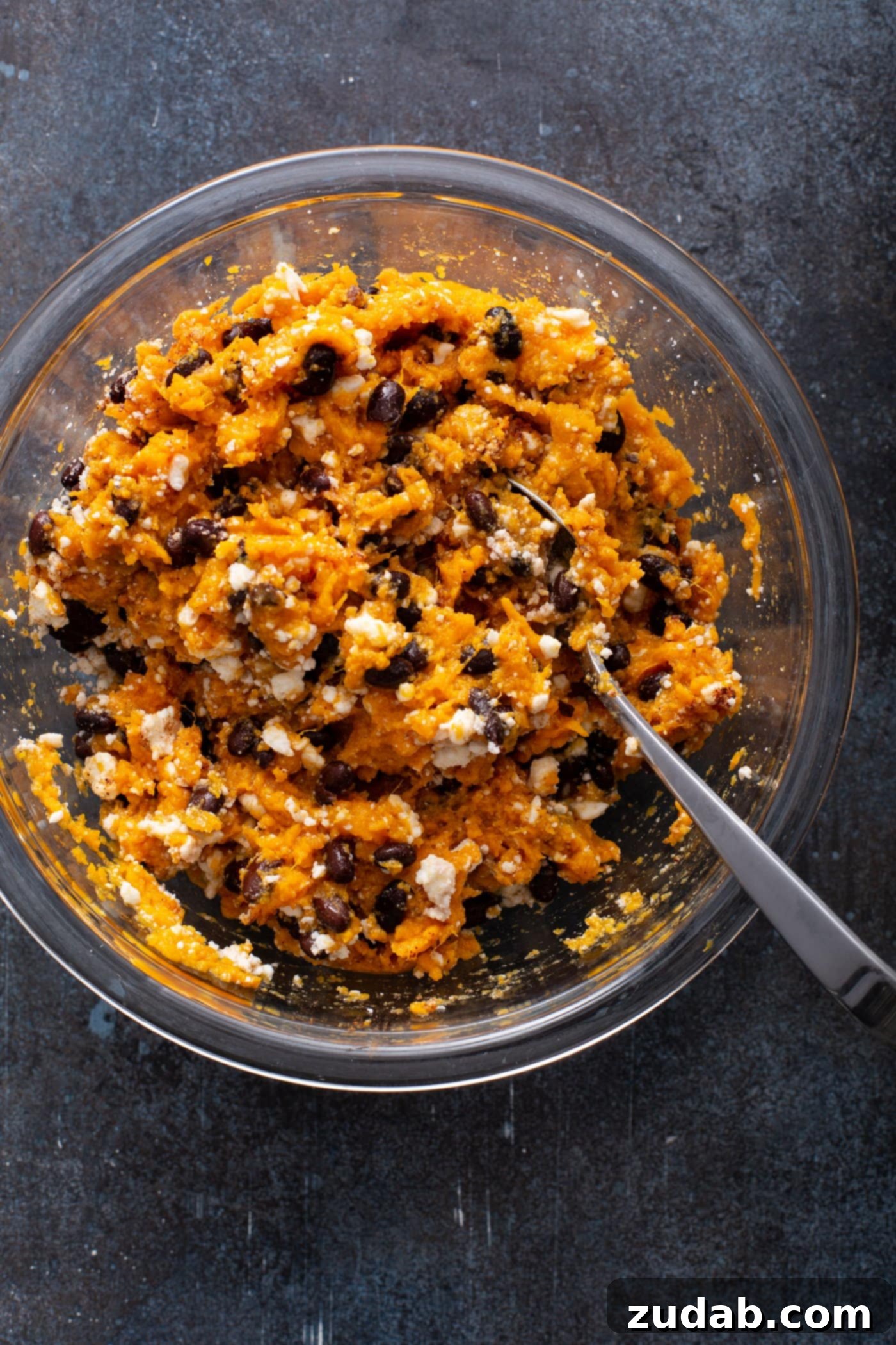 A glass bowl with a mixture of mashed sweet potato, black beans, crumbled queso fresco cheese, chili powder, and cinnamon. A spoon is resting in the mixture, ready for the next step.