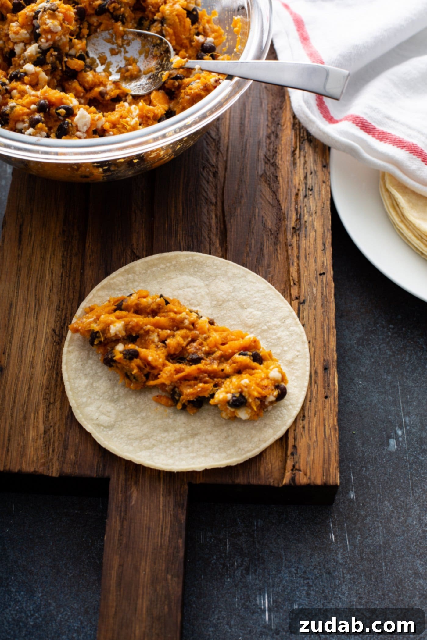 Sweet potato filling with black beans and queso fresco cheese on an open corn tortilla, ready to be rolled. There is a bowl of enchilada filling and a plate of corn tortillas covered with a red and white kitchen towel in the background, illustrating the assembly process.