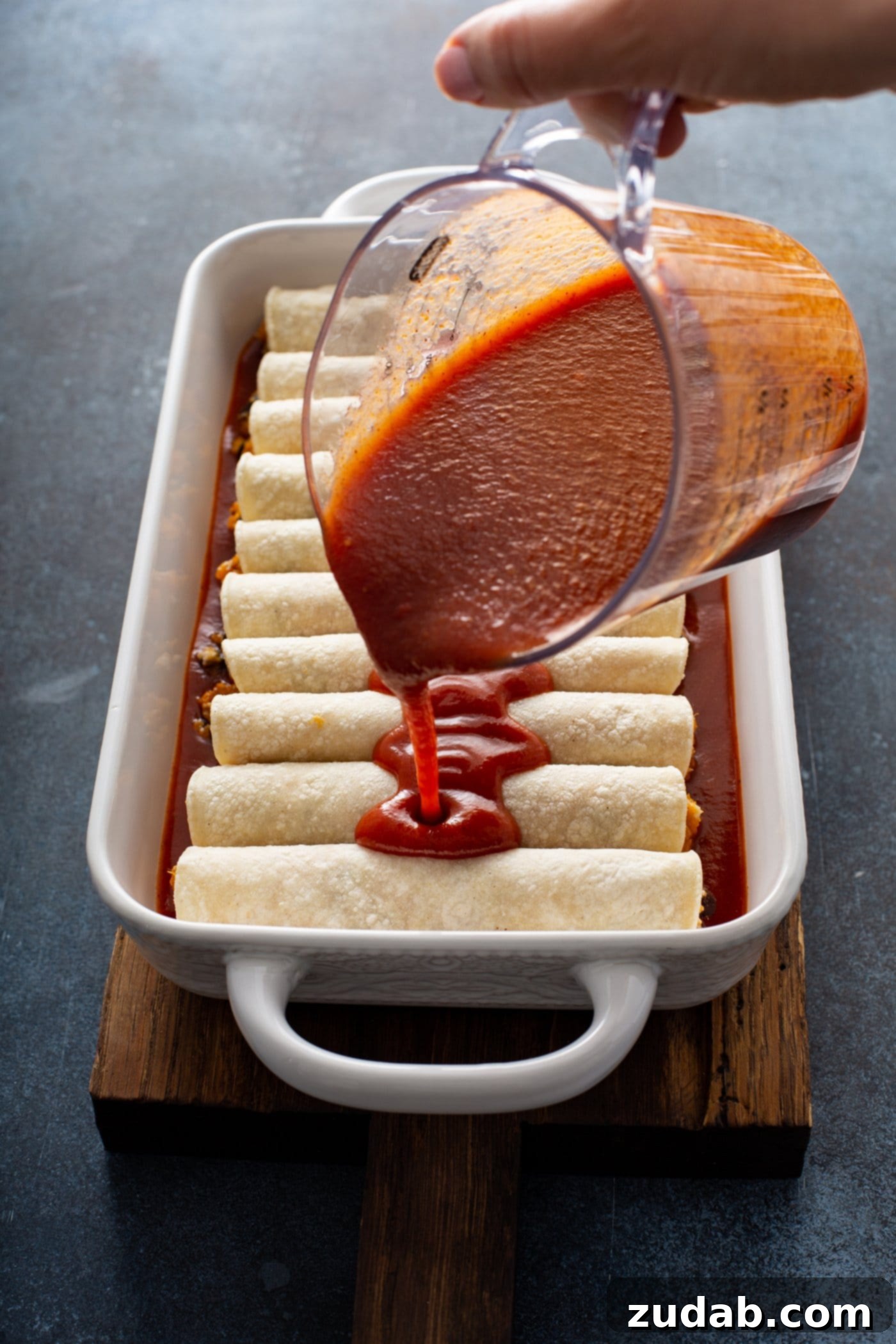 Unbaked enchiladas arranged in a white baking dish. Red enchilada sauce is being poured from a glass measuring cup over the rolled enchiladas, preparing them for baking.