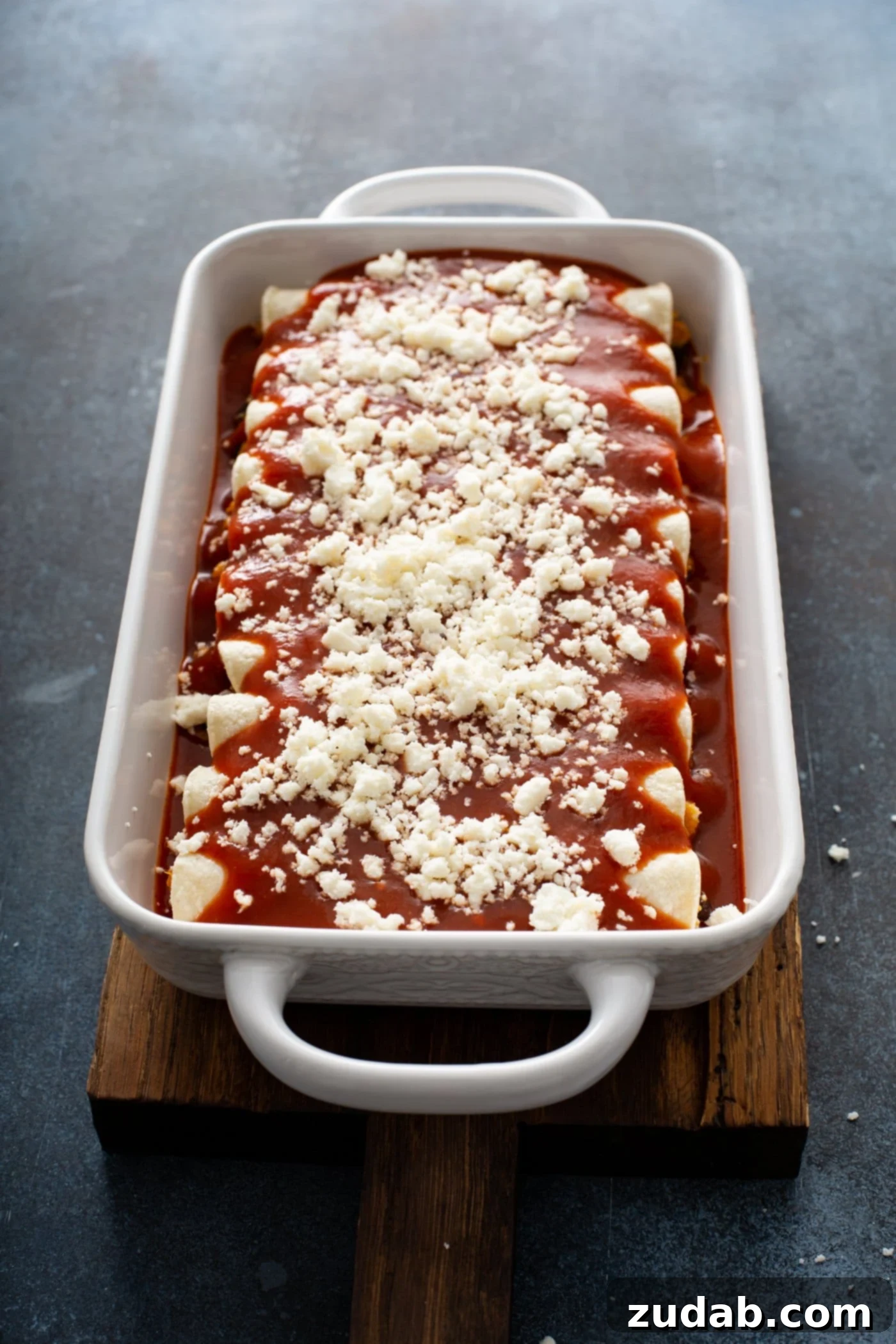 A casserole dish filled with sweet potato enchiladas, freshly baked. They are topped with a vibrant red enchilada sauce and a generous sprinkle of melted queso fresco cheese, bubbling at the edges.