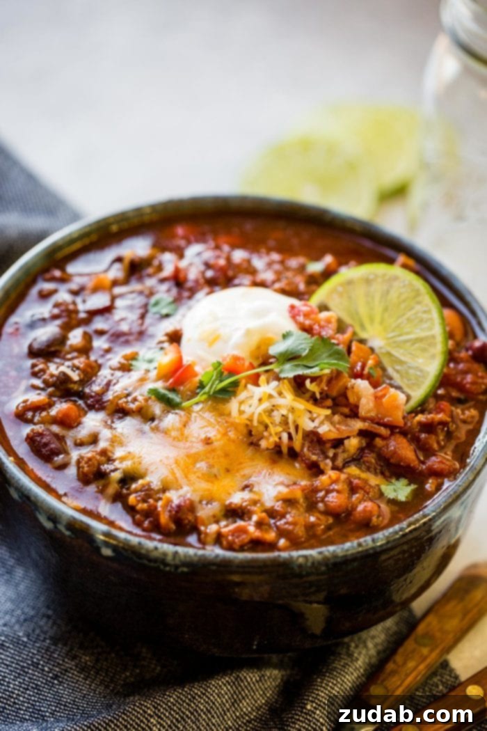 a photo of a bowl full of hearty chili topped with melting cheddar cheese, fresh cilantro, bacon crumbs, a slice of lime and a scoop of sour cream.