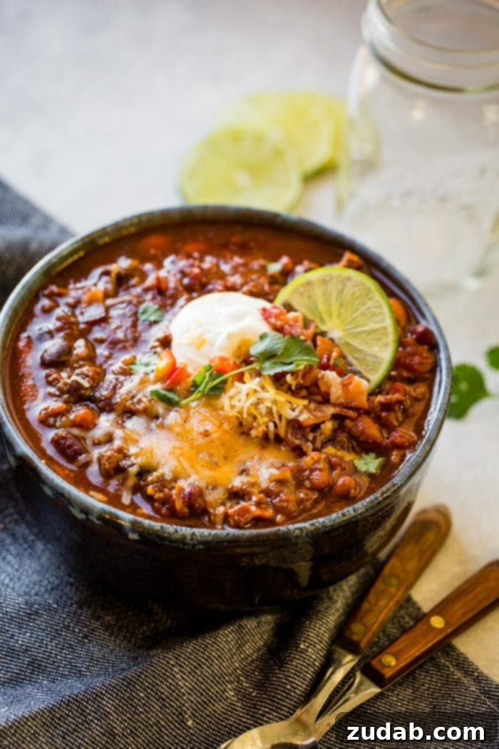 a photo of a bowl full of classic chili loaded with beans, ground beef, and bacon and topped with shredded cheese, sour cream, cilantro and a slice of lime.