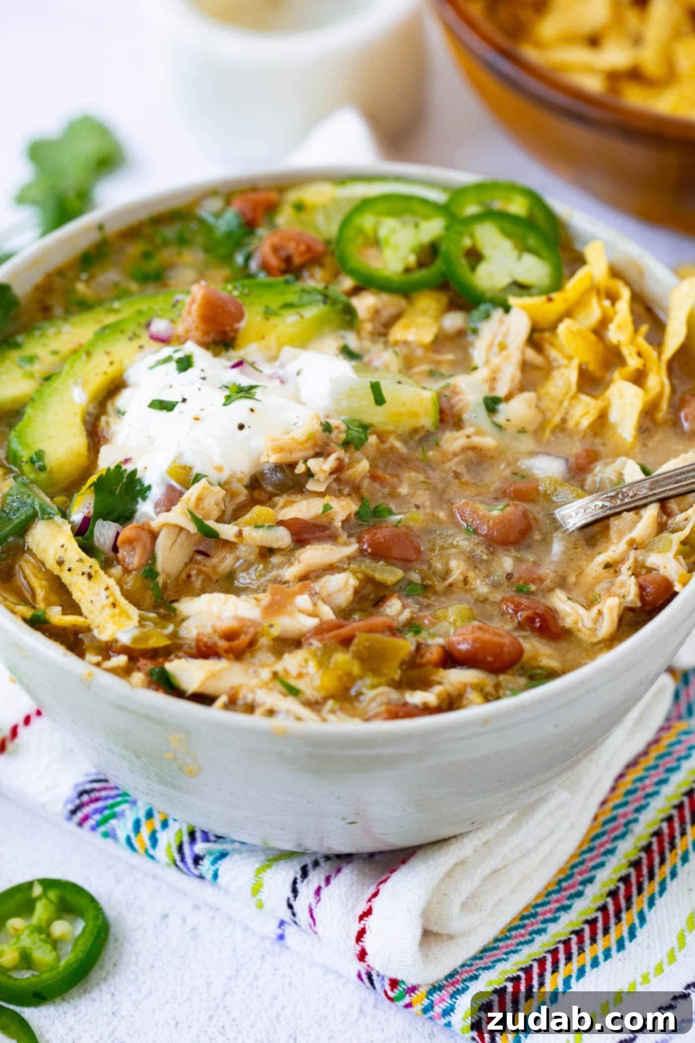 A close-up of a creamy white chicken chili bowl, garnished with sour cream, avocado, tortilla strips, and jalapenos, with a spoon.