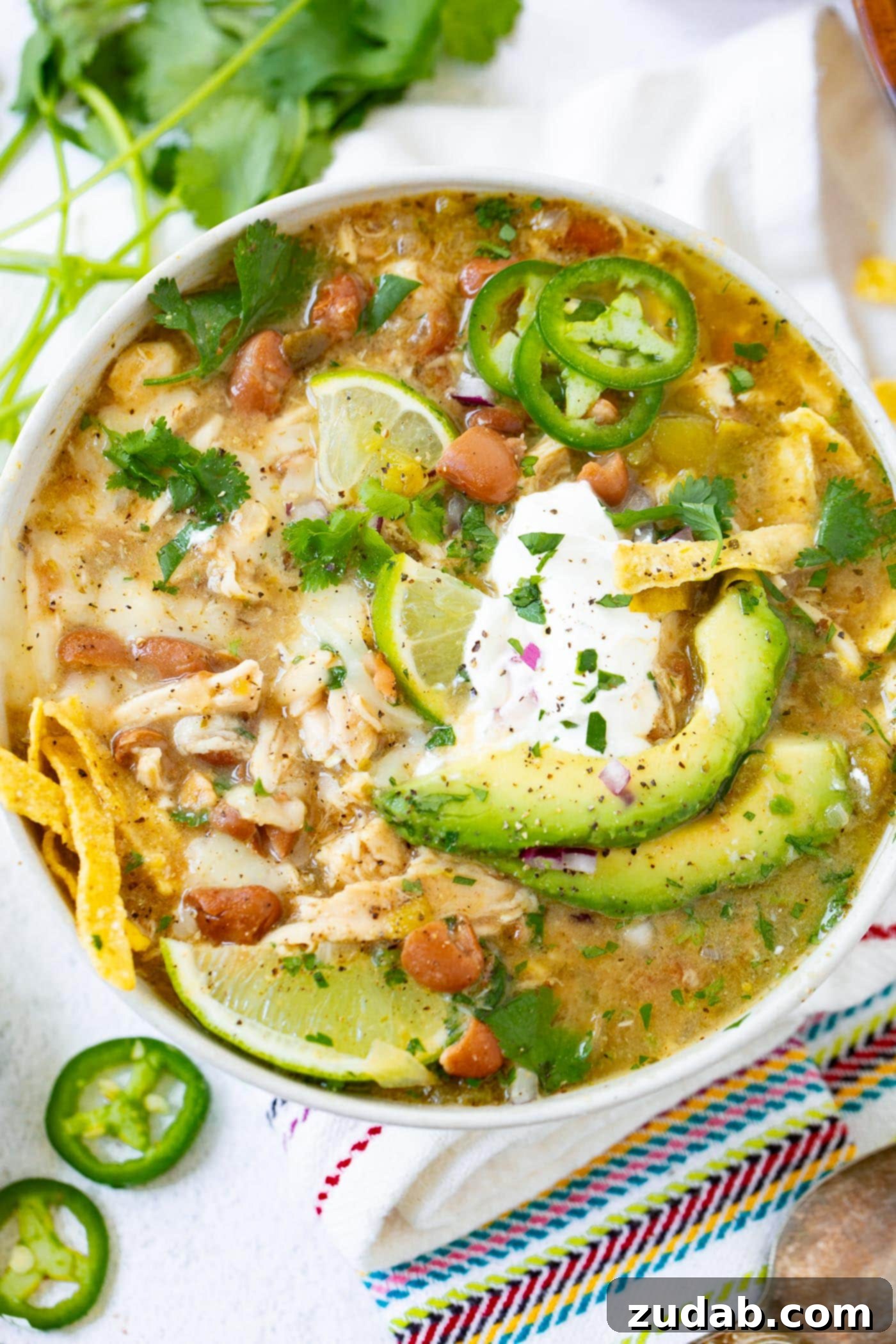 Appetizing white chicken chili in a bowl, generously topped with sour cream, sliced avocado, crunchy tortilla strips, and fresh jalapenos.