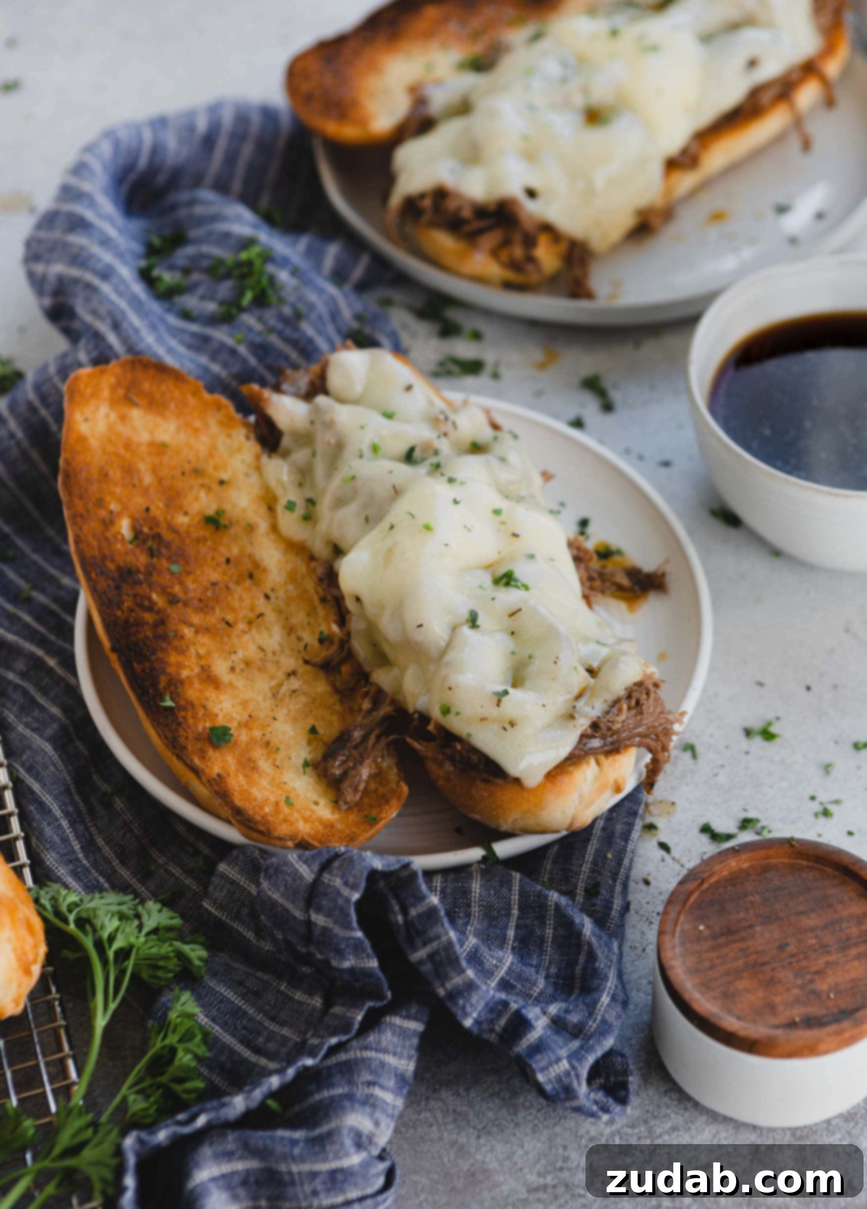 Ultimate Slow Cooker French Dip Sandwiches 2 An opened French dip sandwich on a plate with melted Swiss cheese and tender beef. A bowl of rich au jus is beside it, with another sandwich in the background.