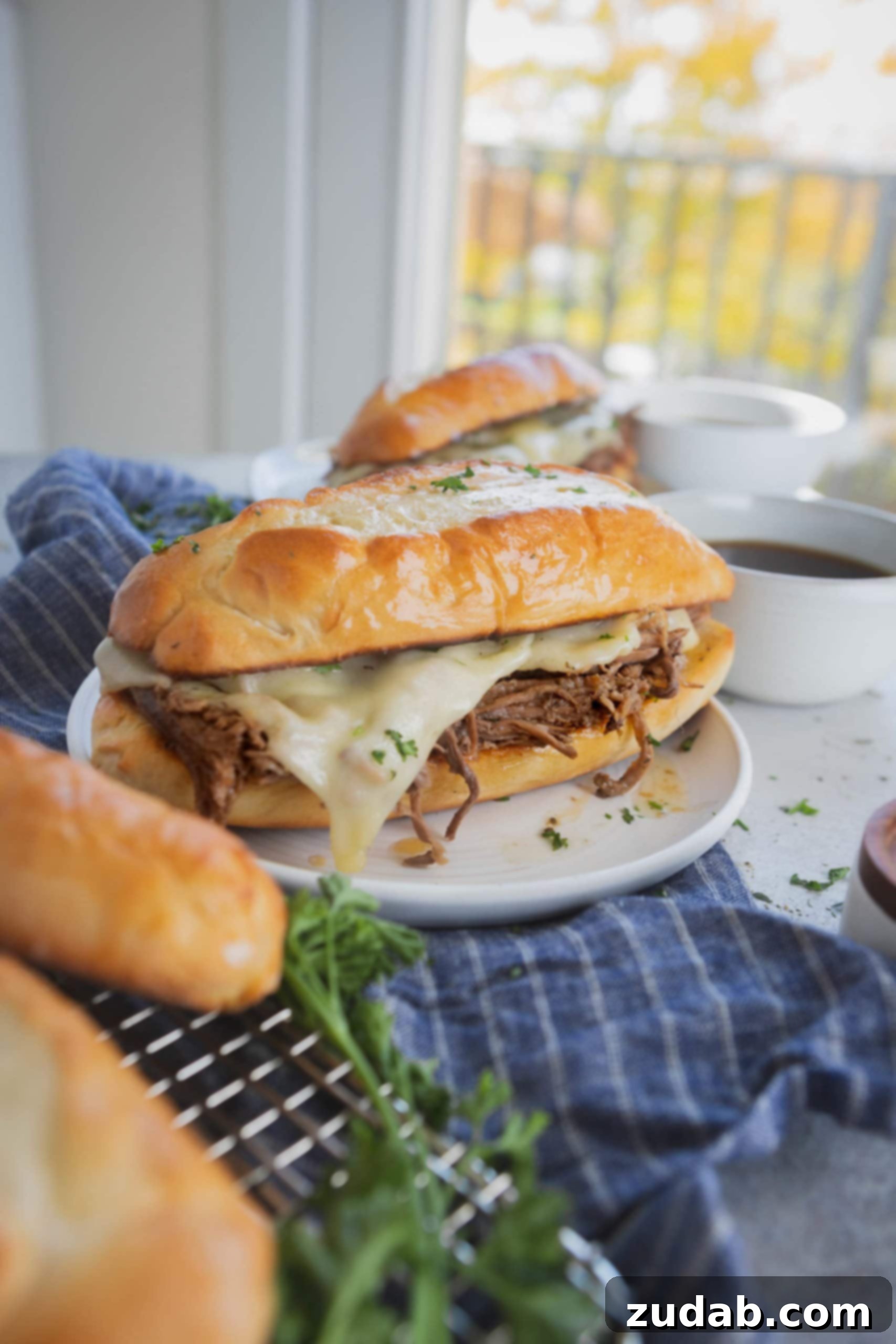 Ultimate Slow Cooker French Dip Sandwiches 4 A perfectly toasted French dip sandwich with melted Swiss cheese and shredded beef on a white plate. Two more hoagie rolls and a sandwich are visible in the background.