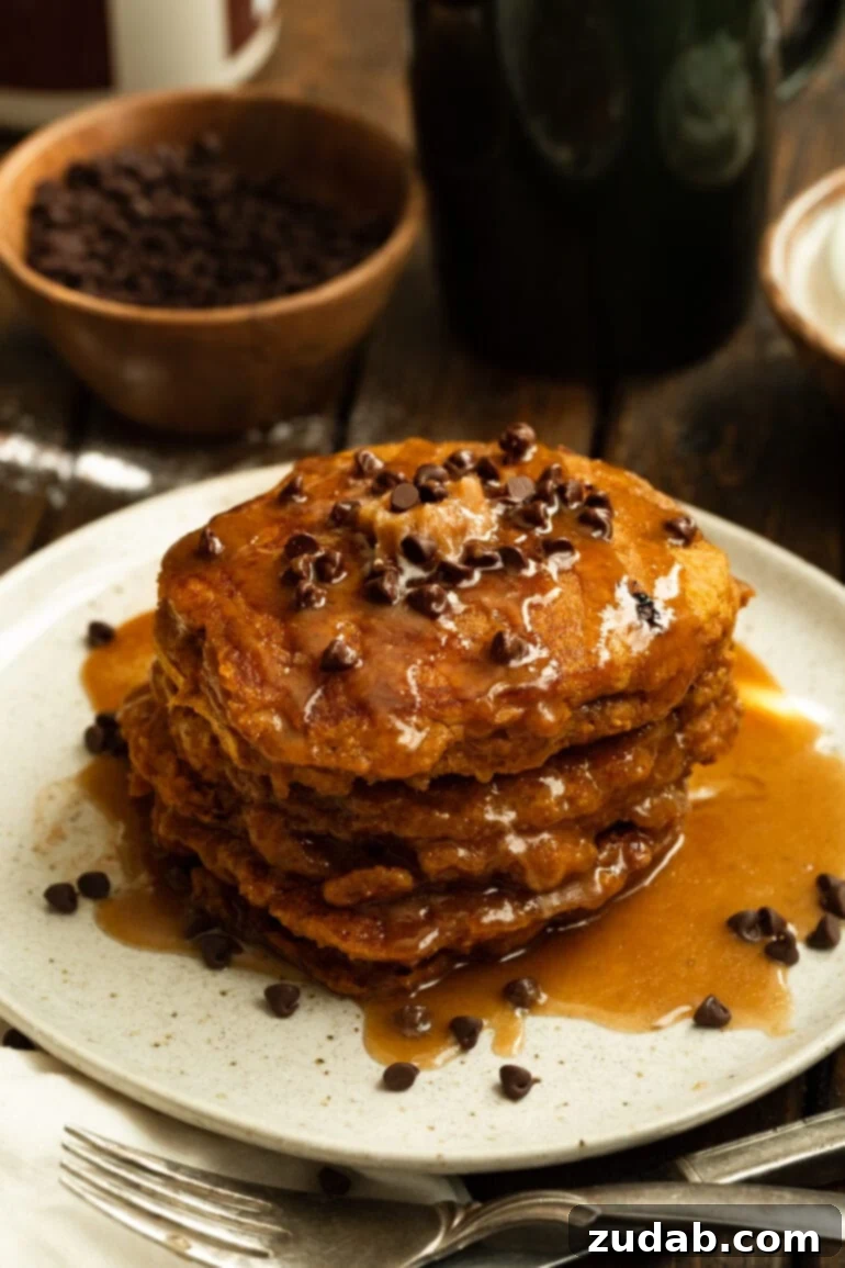 Homemade Whole Wheat Pumpkin Pancakes with Cinnamon Caramel Syrup 6 A beautifully composed stack of whole wheat pumpkin pancakes, generously coated in cinnamon caramel syrup and adorned with a sprinkle of mini chocolate chips, on a white plate.