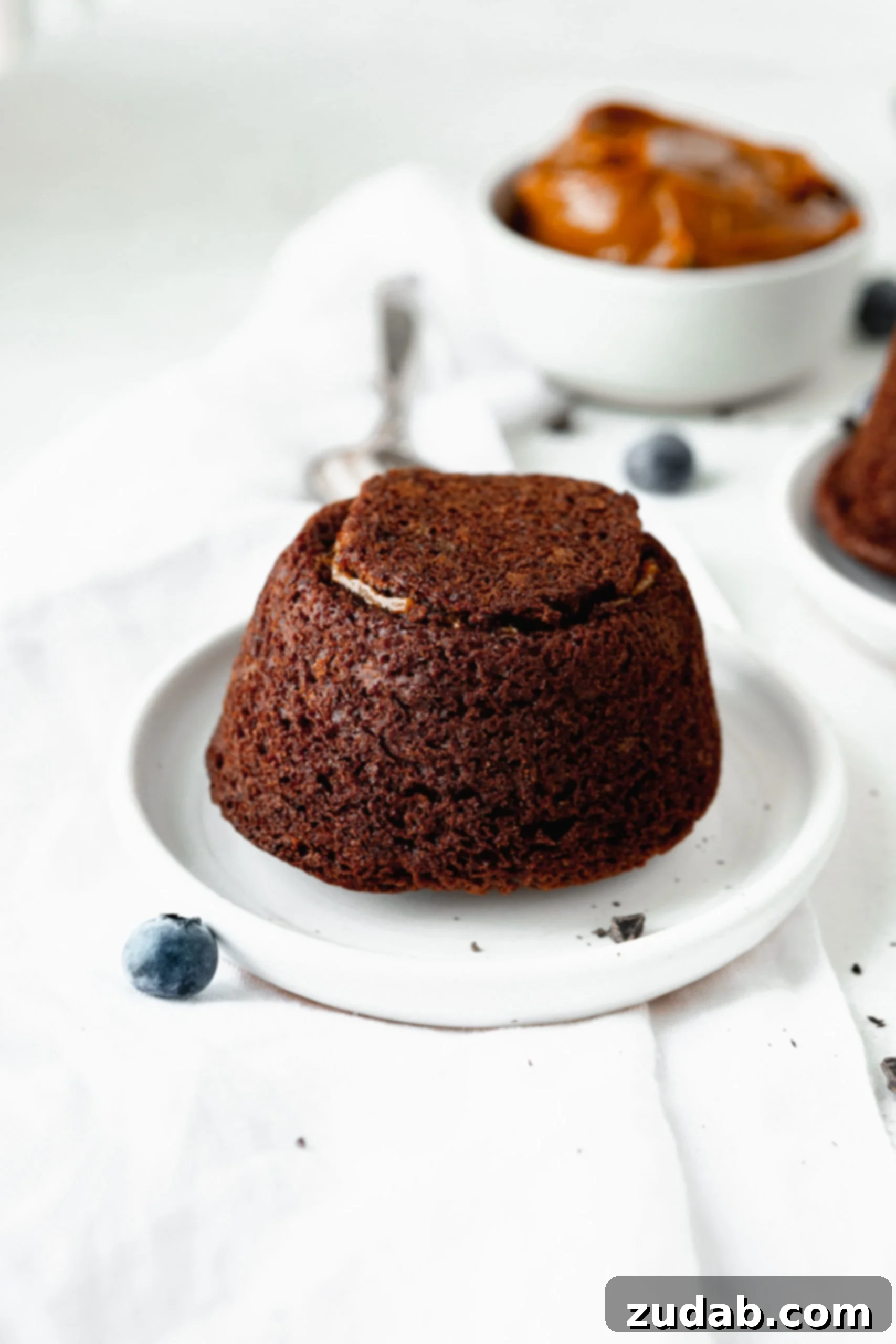 Chocolate Caramel Molten Lava Cakes A chocolate molten lava cake on a white dessert plate. There are a few blue berries scattered around and a bowl of caramel in the background.