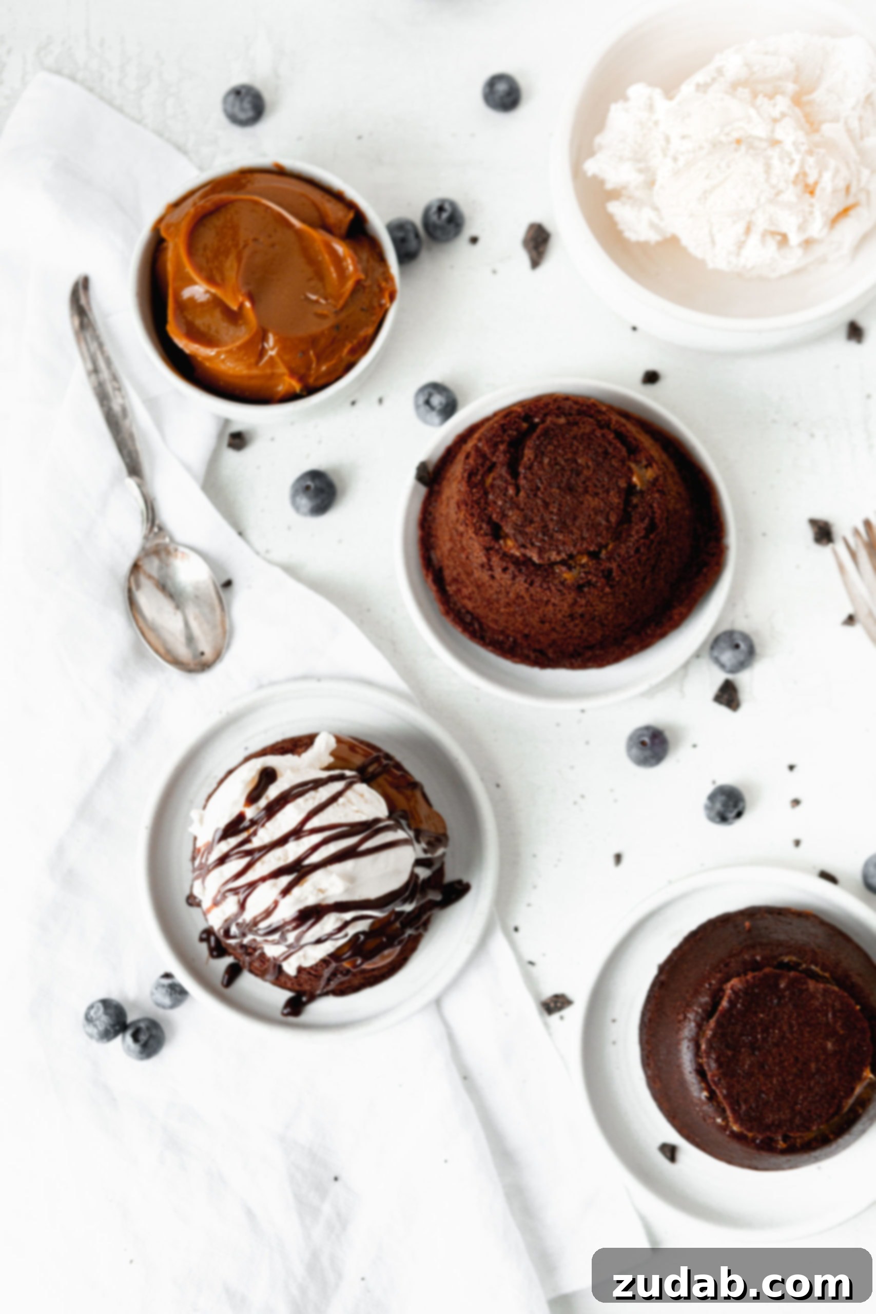Chocolate Caramel Molten Lava Cakes Molten lava cakes on dessert plates. One has been topped with a scoop of ice cream and chocolate drizzles. There are a few blue berries scattered on the table and a spoon is next to a bowl of caramel.