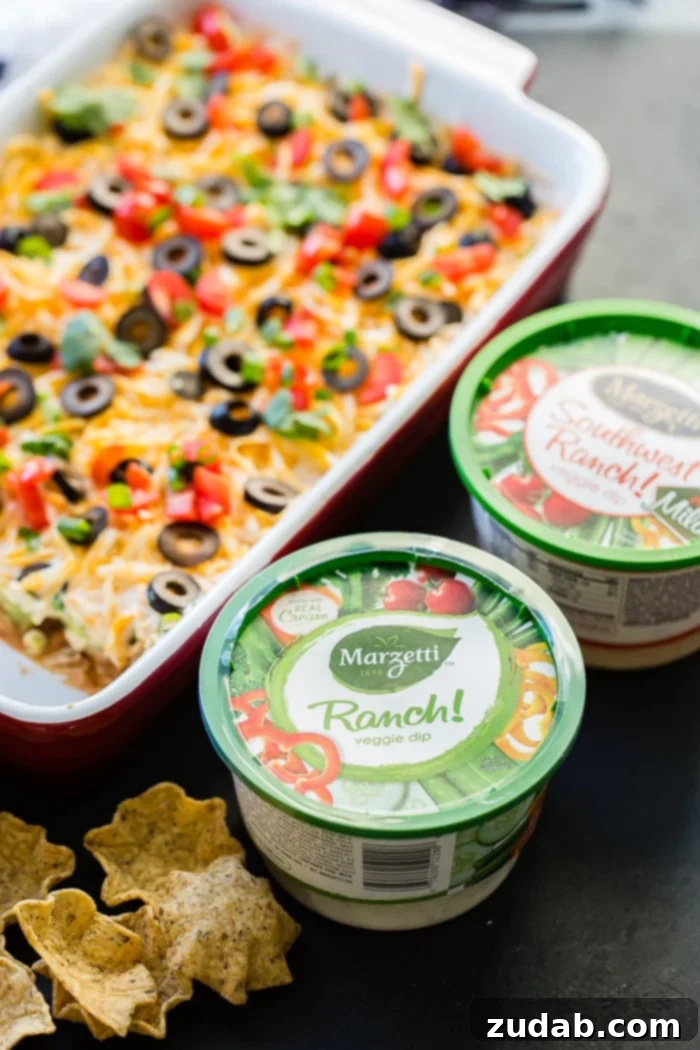 Ultimate Fiesta 7-Layer Dip 2 A vibrant Mexican 7 layer dip, perfectly layered with refried beans, guacamole, Southwest ranch, cheese, olives, tomatoes, and green onions, served in a clear dish with tortilla chips ready for dipping.