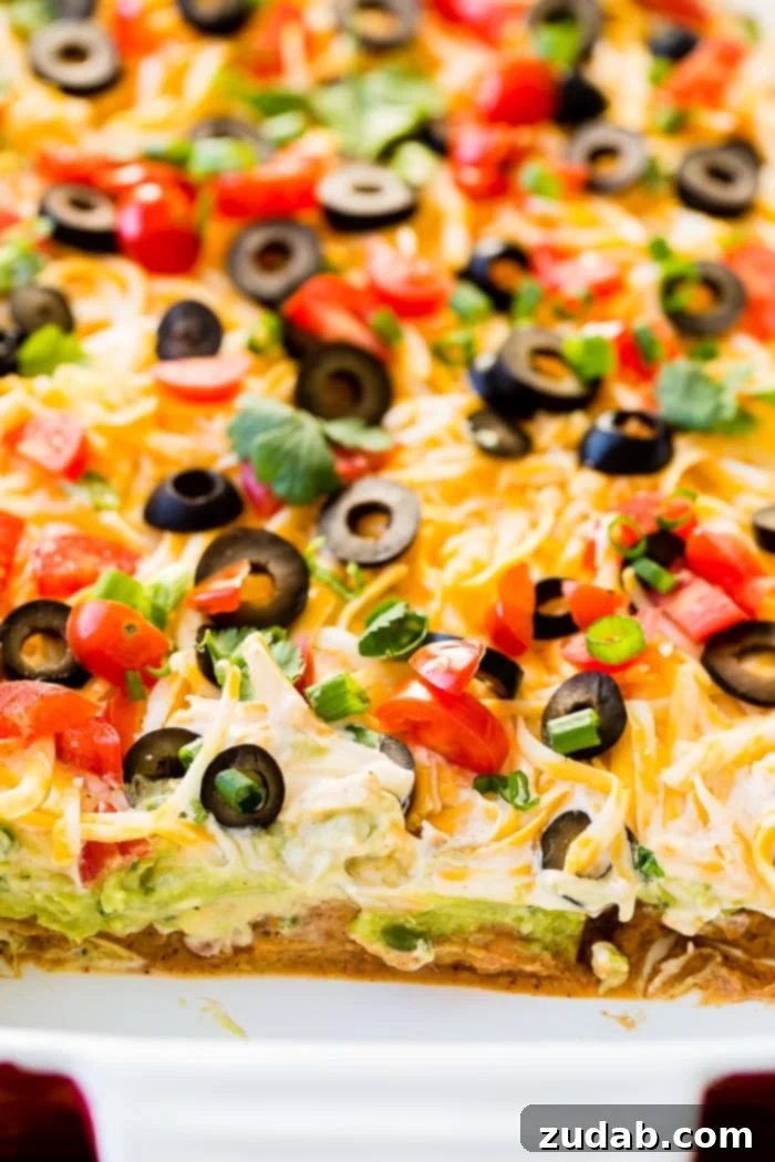 Ultimate Fiesta 7-Layer Dip 4 A close-up shot of a large baking dish filled with Mexican 7 Layer Dip, showcasing the distinct layers of beans, guacamole, ranch, cheese, olives, and tomatoes, ready to be served.