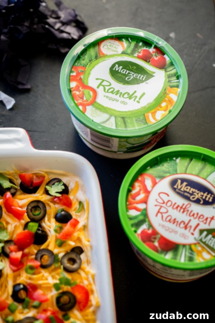 Ultimate Fiesta 7-Layer Dip 5 Two tubs of creamy Marzetti ranch dressing stand next to a freshly prepared pan of vibrant Mexican 7 layer dip, highlighting the secret ingredient for this delicious recipe.