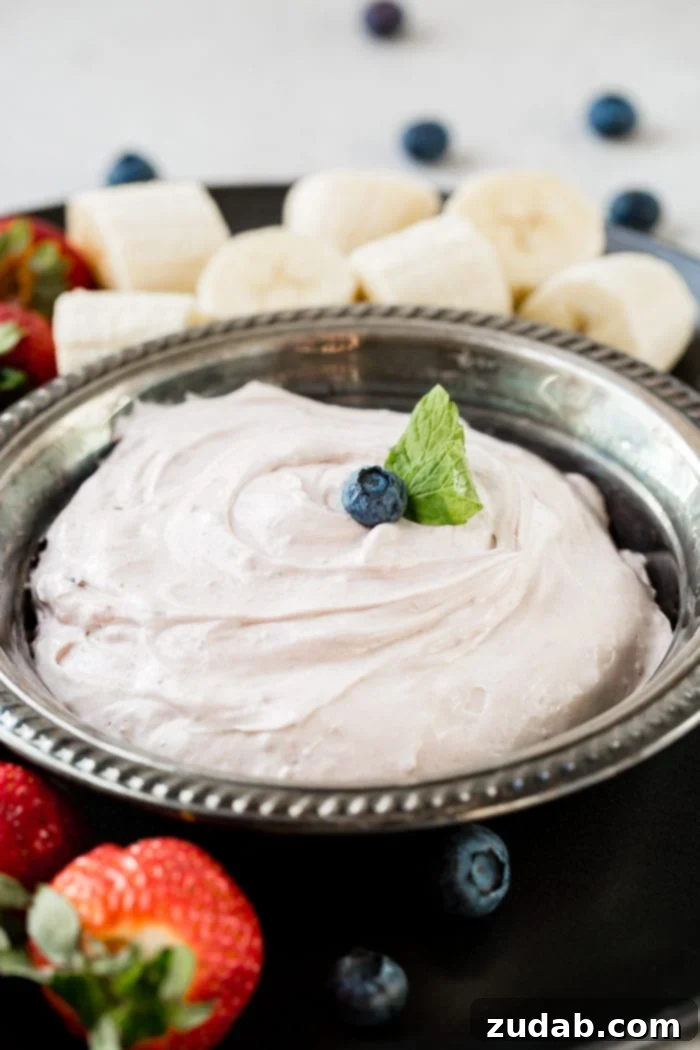 Blueberry Cloud Fruit Dip 2 A beautifully presented silver bowl filled with creamy Blueberry Fluff Fruit Dip, surrounded by a colorful assortment of fresh fruits and crunchy dippers, inviting everyone to enjoy a delightful spring treat. The vibrant blueberries and lush strawberries suggest a refreshing and easy-to-make dessert, perfect for any occasion.