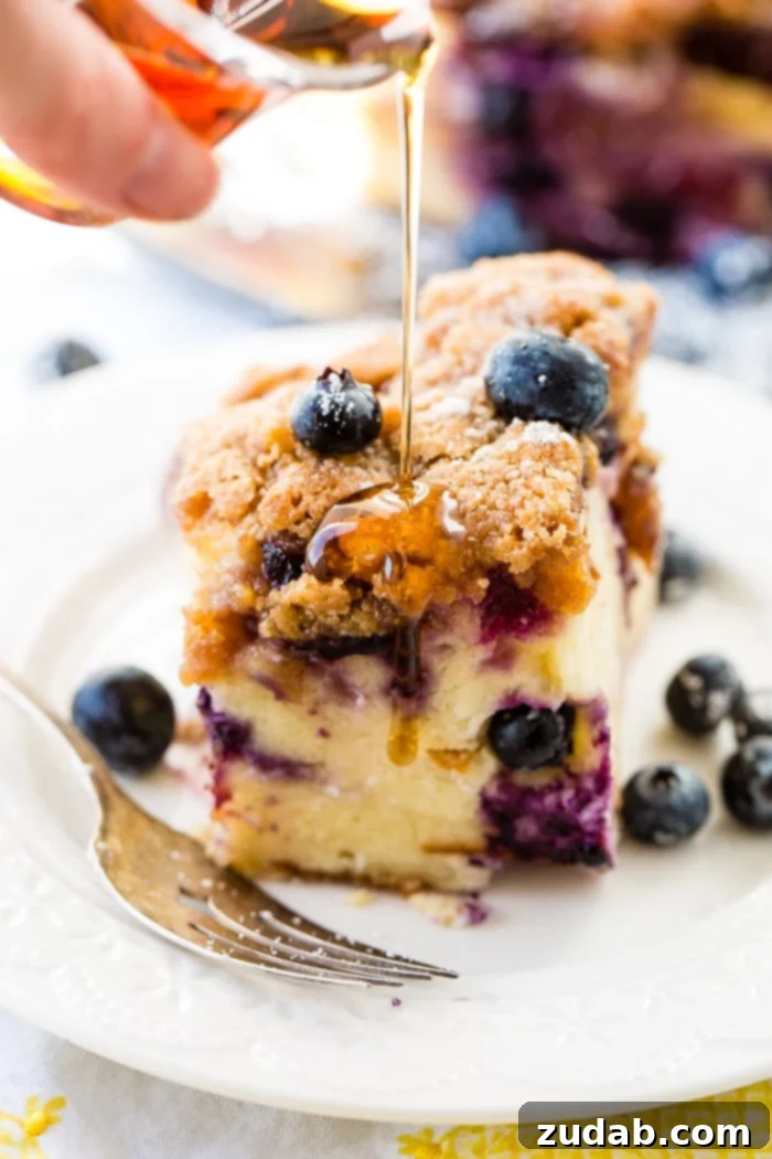 3 Irresistible Instant Pot Blueberry French Toast Bakes 4 A square slice of French toast casserole, garnished with fresh blueberries and powdered sugar, with maple syrup being drizzled over the top. Served on a white lace plate with a vintage fork, with the Instant Pot in the background.