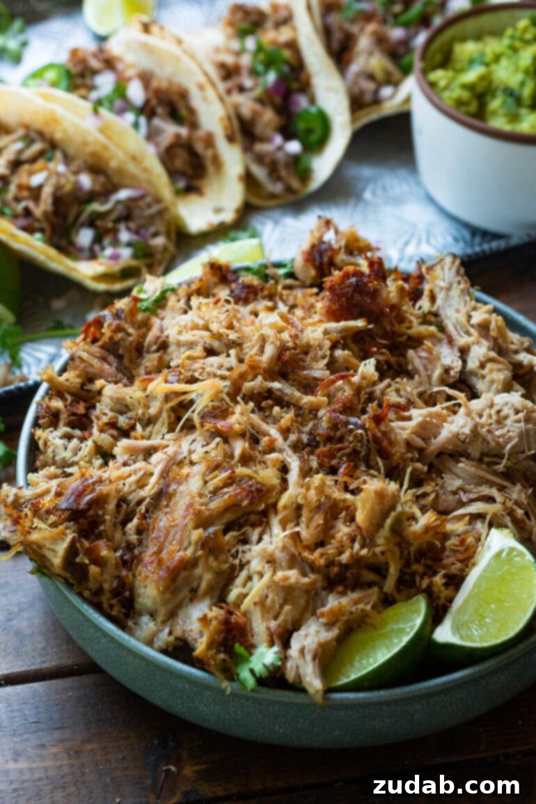 Crockpot Carnitas: Crispy & Juicy Pulled Pork 2 A beautifully arranged bowl of shredded pork carnitas, garnished with fresh cilantro and lime wedges, with three loaded tacos blurring in the background.