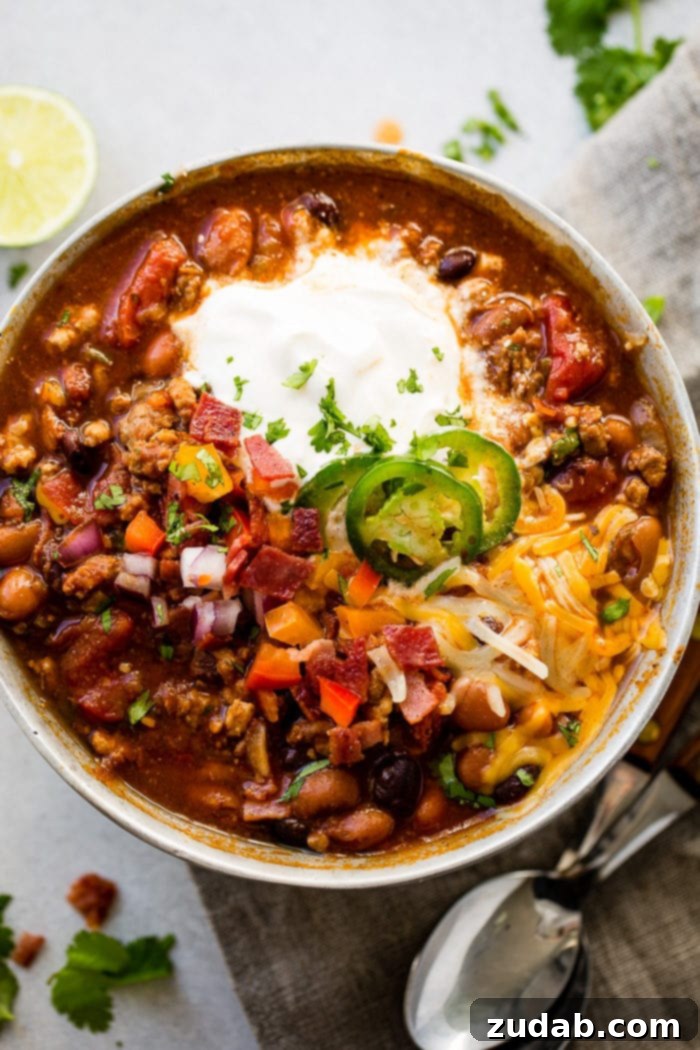 Prize-Winning Lean Instant Pot Turkey Chili 2 A metal bowl of ground turkey chili full of beans, red onions that have been chopped, chopped peppers, jalapeno slices on melted cheese next to a dollop of sour cream with a dash of cilantro