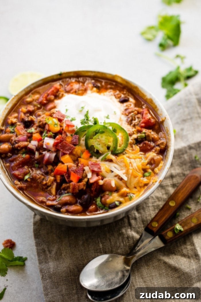 Prize-Winning Lean Instant Pot Turkey Chili 3 A metal bowl of ground turkey chili full of beans, red onions that have been chopped, chopped peppers, jalapeno slices on melted cheese next to a dollop of sour cream with a dash of cilantro and a grey napkin underneath with two wooden handled spoons to the side.