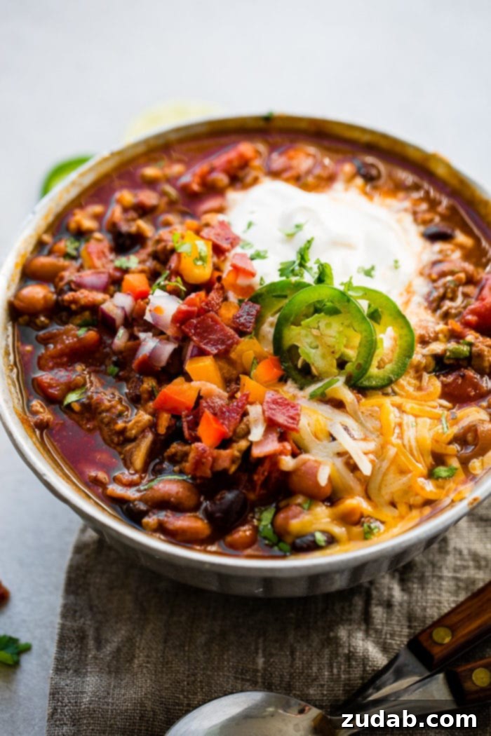 Prize-Winning Lean Instant Pot Turkey Chili 4 A metal bowl of instant pot turkey chili full of beans, red onions that have been chopped, chopped peppers, jalapeno slices on melted cheese next to a dollop of sour cream with a dash of cilantro and a grey napkin underneath with two wooden handled spoons to the side.
