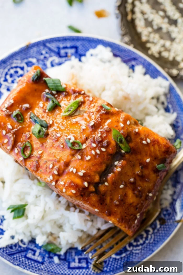 A beautifully glazed Asian sticky salmon fillet, garnished with fresh green onions and sesame seeds, served atop a bed of fluffy white rice in an elegant blue Asian-printed dish.