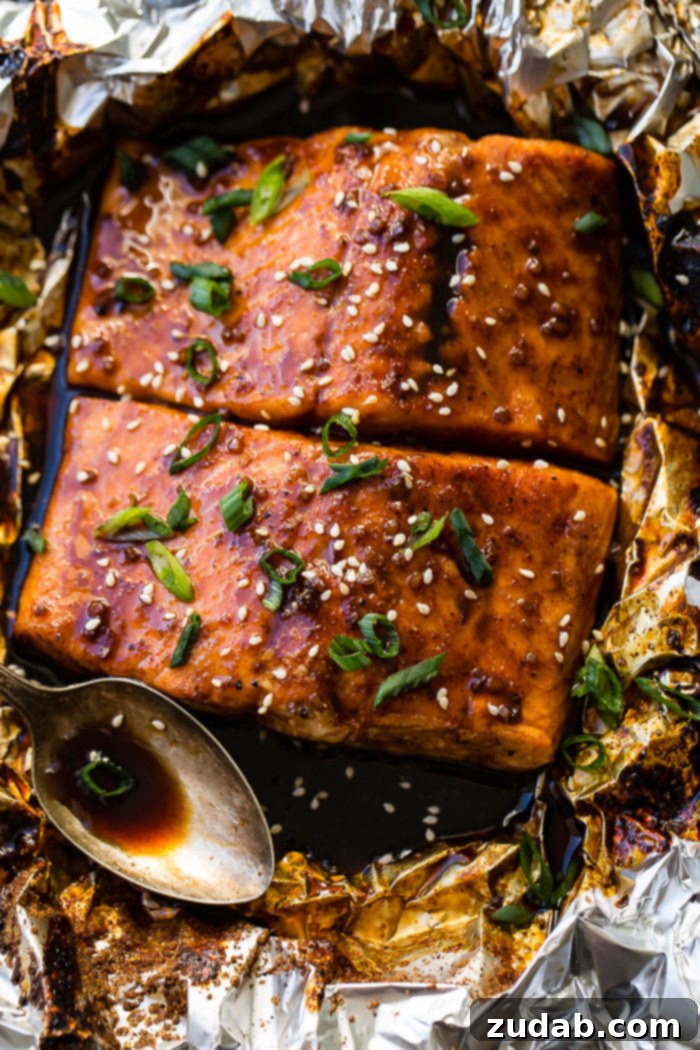 Two raw salmon fillets covered in a rich, dark Asian sticky sauce with green onions and sesame seeds, carefully wrapped in foil, ready for baking.