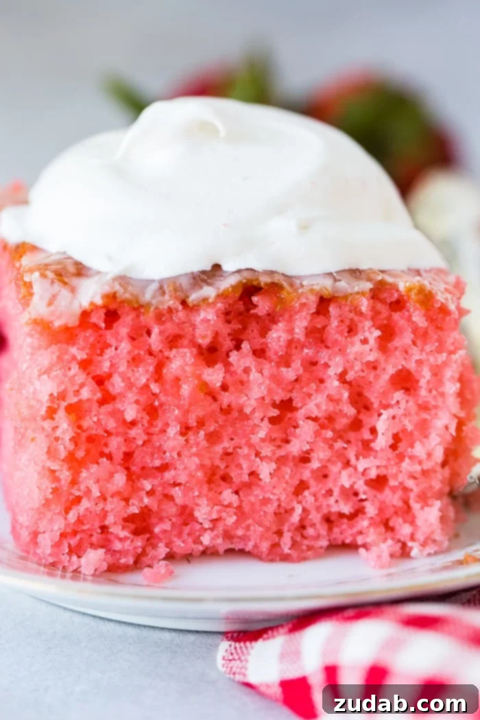 Wobbly Strawberry Dream Cake 2 A china plate with a large slice of bright pink strawberries and cream jello cake with a dollop of whipped cream on top with a red and white checkered napkin underneath.