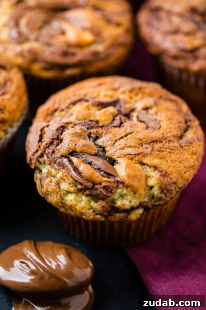 Nutella-Marbled Banana Muffins 2 A close-up photograph of a moist Nutella swirled banana muffin with more delicious muffins blurred in the background, ready to be enjoyed for breakfast or brunch.