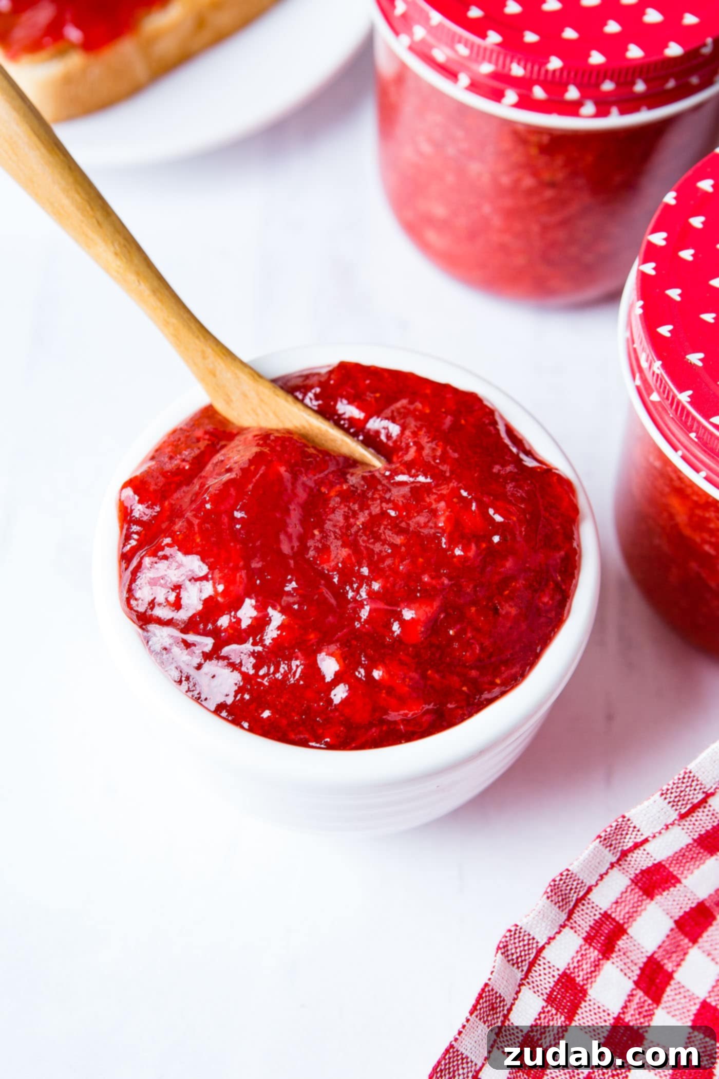 A white bowl with strawberry jam in it. There is a small wooden spoon in the jam, and jars of jam in the background.