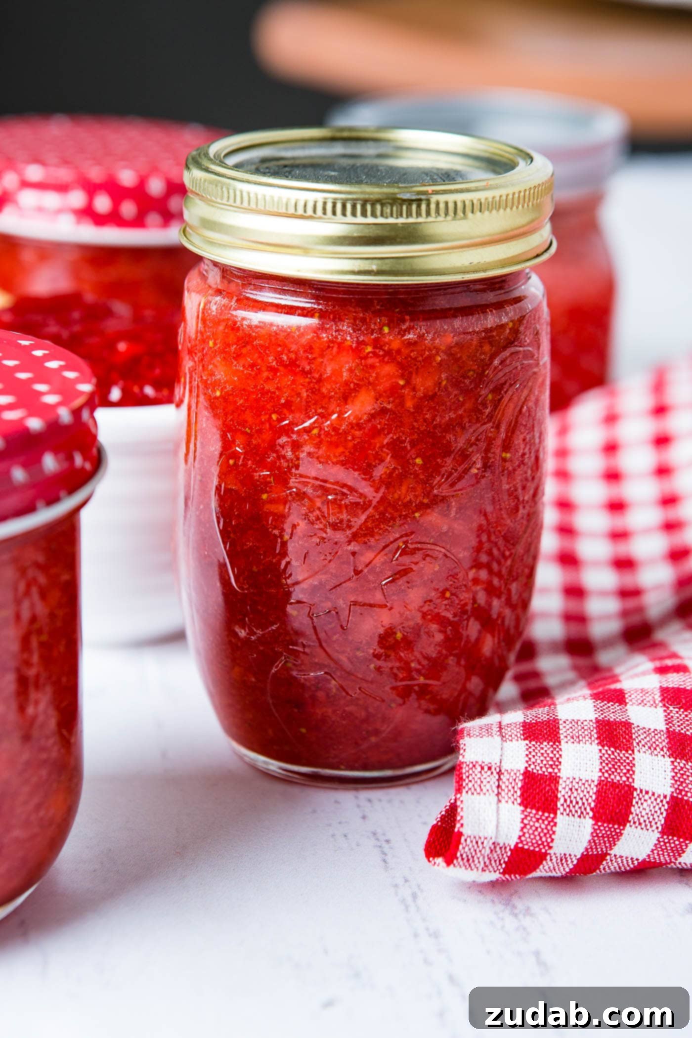 Four jars of homemade strawberry jam. There are red and white checked lids on some of the jars and a red and white checked tea towel next to the jars of jam.