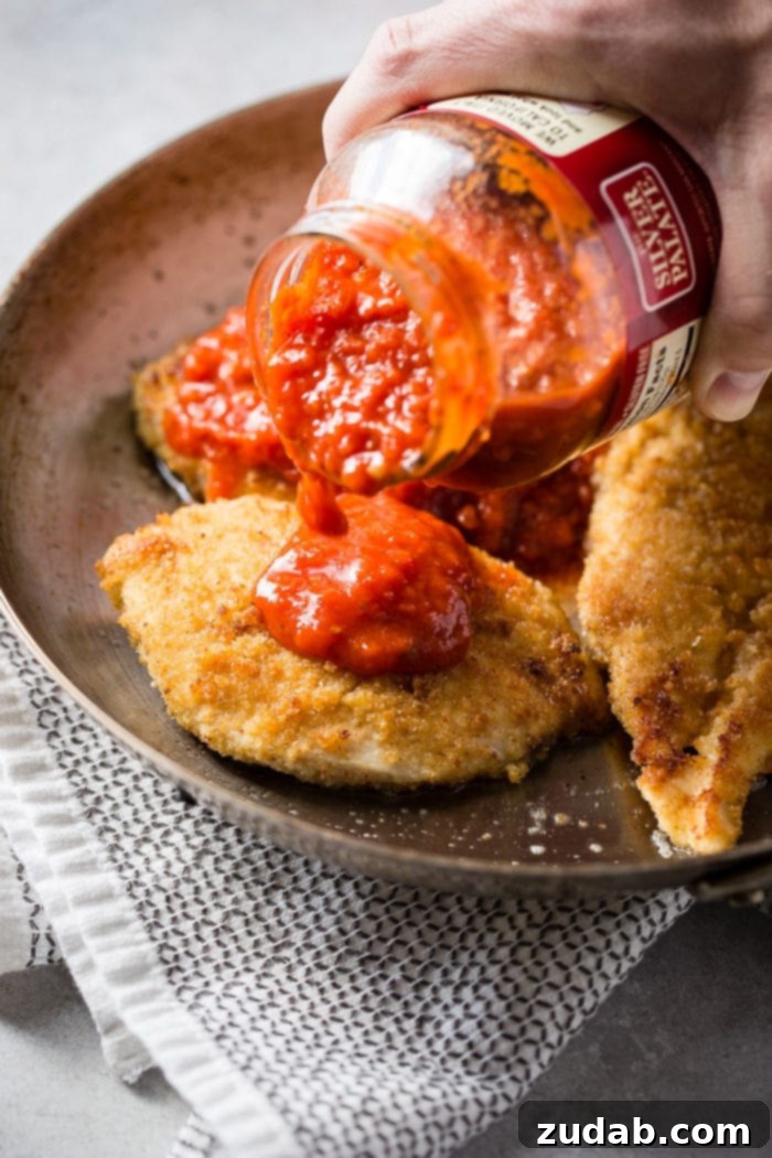 Speedy Weeknight Chicken Parmesan 3 Golden brown breaded chicken cutlets sizzling in a cast-iron skillet, with a jar of rich marinara sauce being poured generously over each piece. This is the initial step for a quick and easy shortcut chicken parmesan using herbed croutons for the breading.