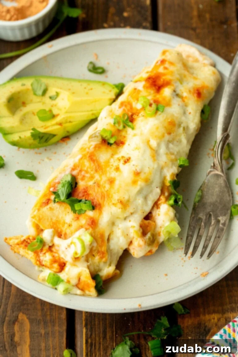 Simple and Delicious Cheesy Chicken Enchiladas 6 a photo of a cheesy, creamy chicken enchilada in a flour tortillas sitting on a dinner plate with a fork next to it and some sliced avocado