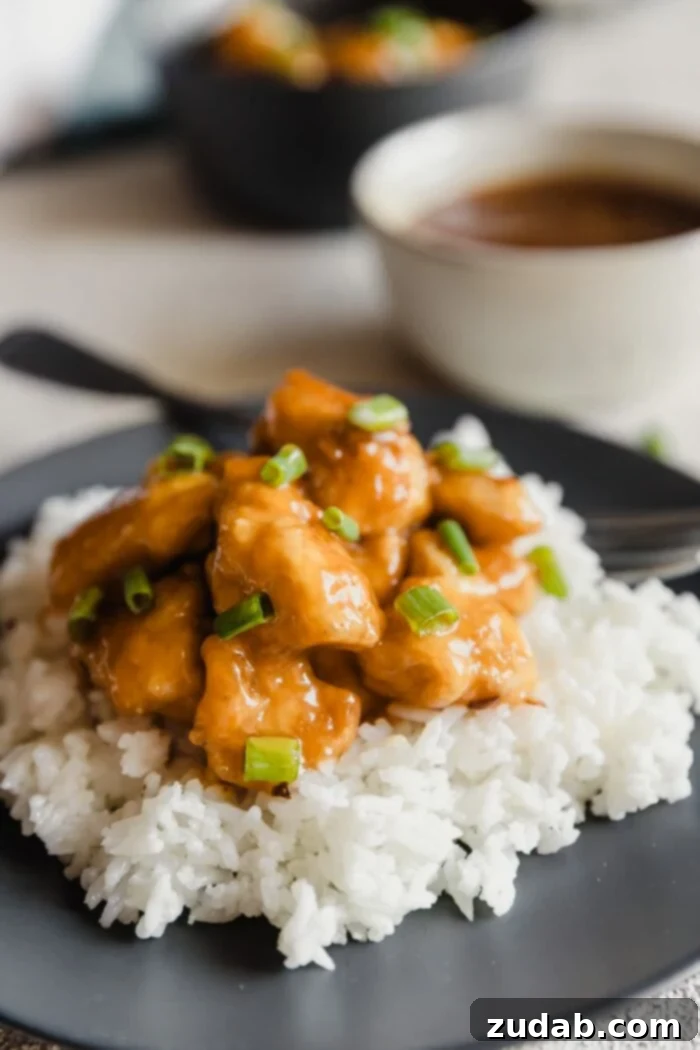 A dark plate with fluffy white rice and orange chicken sprinkled with green onions over the top and a white bowl with extra orange sauce in the background