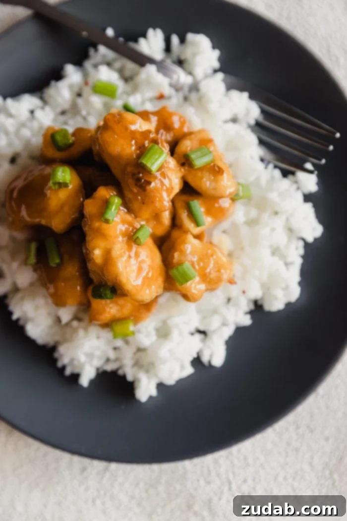 A dark plate with fluffy white rice and orange chicken sprinkled with green onions over the top and a white bowl with extra orange sauce in the background