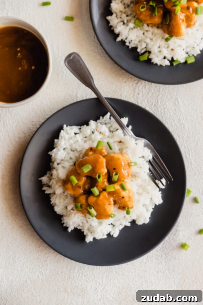 A dark plate with fluffy white rice and orange chicken sprinkled with green onions over the top and a white bowl with extra orange sauce in the background
