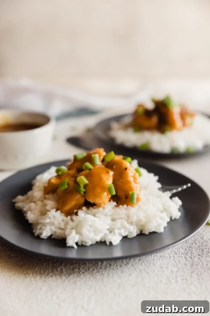 A dark plate with fluffy white rice and orange chicken sprinkled with green onions over the top and a white bowl with extra orange sauce in the background