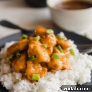A dark plate with fluffy white rice and orange chicken sprinkled with green onions over the top and a white bowl with extra orange sauce in the background