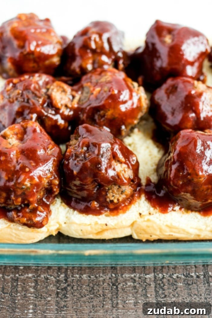 Effortless Smoky BBQ Meatball Sliders 6 Cooked BBQ meatballs arranged on the bottom half of Hawaiian slider buns, ready for the next layers of cheese and toppings in a baking dish.