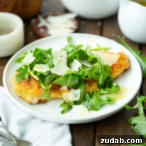 Guilt-Free Chicken Parmesan 2 A square image of a golden breaded chicken breast on a white dinner plate, topped with salad greens, lemon vinaigrette, and shaved Parmesan.