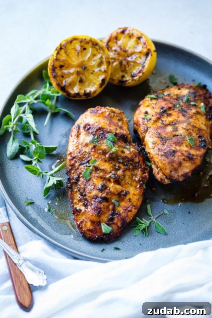 Effortless Oregano Grilled Chicken 4 A grey plate with sliced quick grilled chicken with oregano, fresh oregano sprigs, and grilled lemon halves behind it.