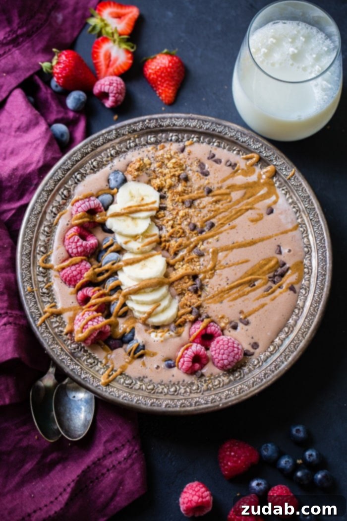 Chocolate Banana Smoothie Bowl Kids Love 2 A vibrant chocolate banana smoothie bowl adorned with fresh raspberries, blueberries, banana slices, crunchy granola, and a drizzle of creamy peanut butter, accompanied by a glass of milk on a rustic wooden board.