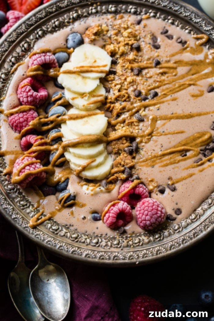 Chocolate Banana Smoothie Bowl Kids Love 3 An overhead shot of a delightful chocolate banana smoothie bowl topped with a colorful array of raspberries, banana slices, blueberries, granola, and chocolate chips, finished with a generous peanut butter drizzle, set against a dark background.