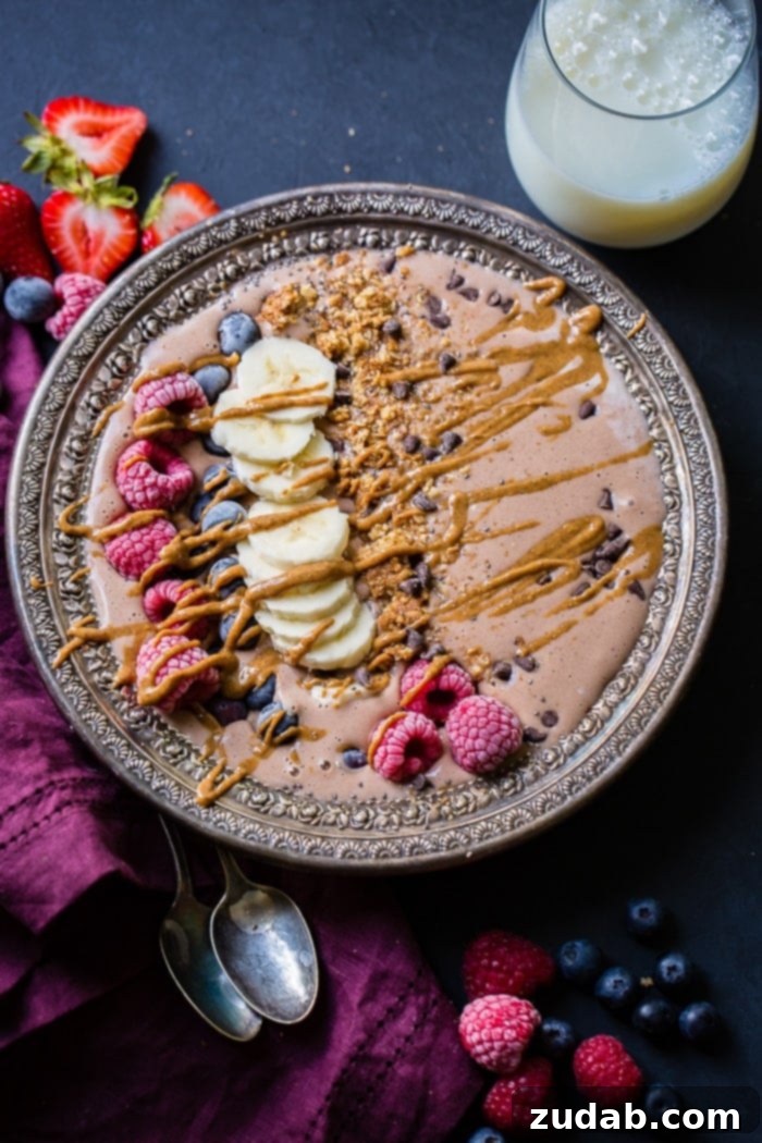 Chocolate Banana Smoothie Bowl Kids Love 5 A beautifully composed chocolate banana smoothie bowl, rich with chocolate chips, fresh raspberries, banana slices, and a lavish drizzle of peanut butter, inviting a healthy and indulgent meal.