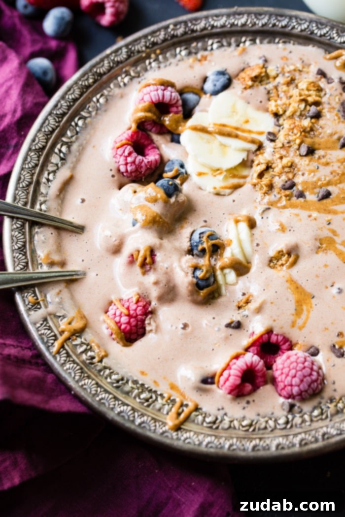 Chocolate Banana Smoothie Bowl Kids Love 8 A stunning overhead view of a fully dressed chocolate banana smoothie bowl, featuring a colorful assortment of raspberries, bananas, blueberries, granola, and chocolate chips, all generously drizzled with peanut butter.