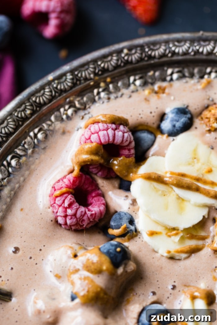 Chocolate Banana Smoothie Bowl Kids Love 9 A sumptuous chocolate banana smoothie bowl, garnished with fresh raspberries, vibrant blueberries, crunchy granola, sweet banana slices, and a rich peanut butter drizzle, ready to be enjoyed.