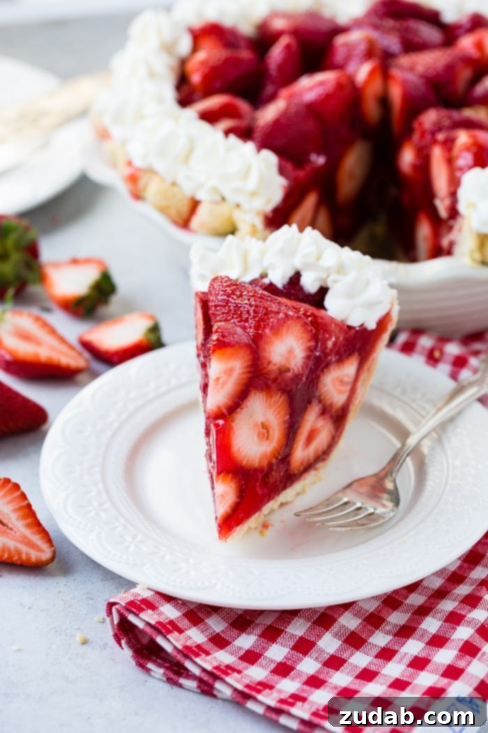 Close-up of Fresh Strawberry Pie Slice A white plate with a big slice of fresh strawberry pie made with jello and a little stabilized whipped cream with a red and white checkered napkin underneath and the whole pie in the background