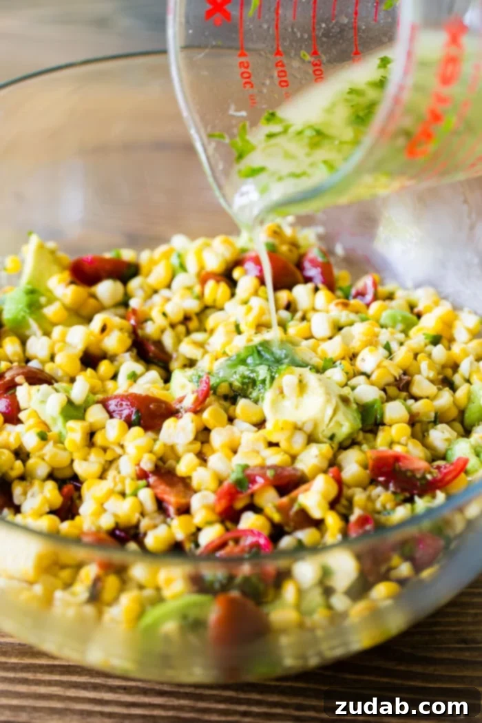 A glistening glass bowl filled with a vibrant grilled corn avocado tomato salad, featuring charred corn kernels, ripe red tomatoes, and creamy avocado chunks. A stream of fresh cilantro lime dressing is being drizzled generously over the top, ready to awaken the flavors.