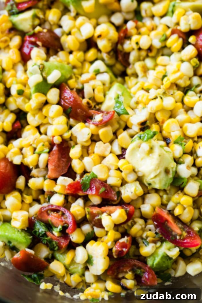 A close-up shot of a fresh Grilled Corn Avocado Tomato Salad, showcasing individual charred corn kernels, juicy halved grape tomatoes, and creamy avocado cubes, all lightly coated in a vibrant green cilantro dressing.
