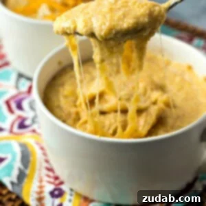 A white bowl full of cheesy grits and a spoon lifting it out of the bowl, showcasing its gooey texture.