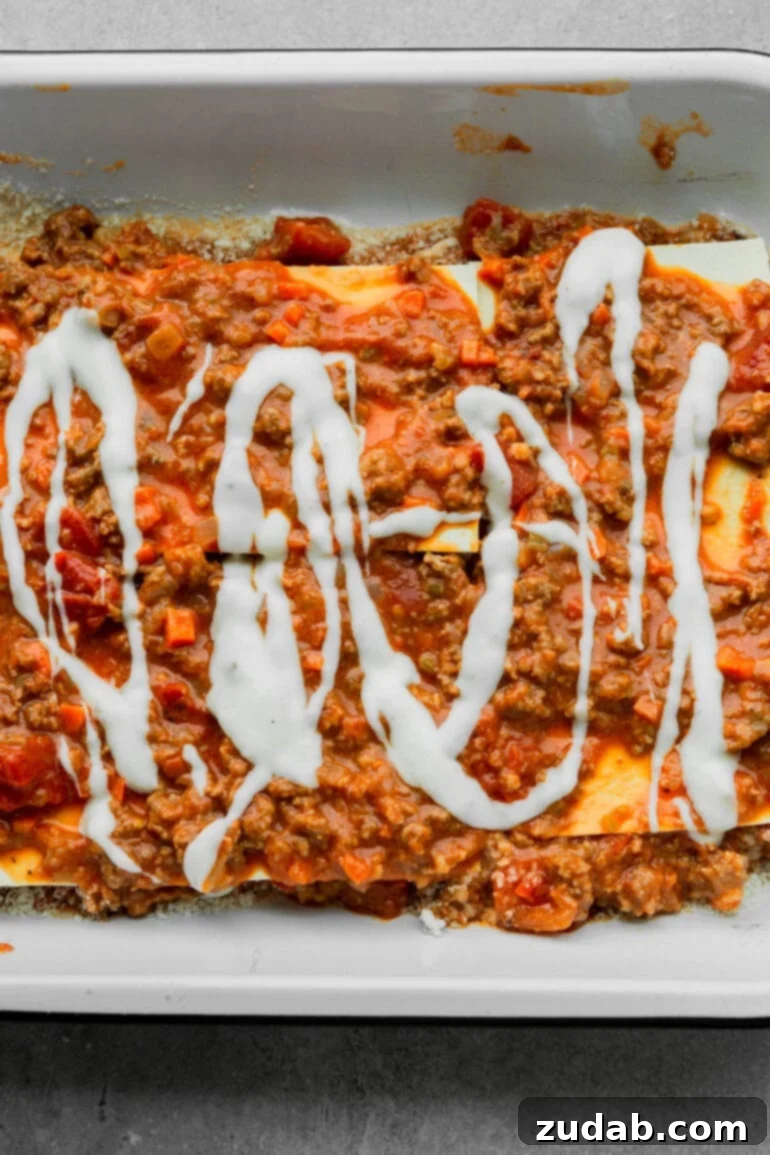 My Go-To Lasagna Masterpiece 7 A captivating overhead shot of a lasagna being carefully assembled in a rectangular baking dish, showing layers of broad pasta noodles, rich meaty sauce, and a generous drizzle of creamy béchamel sauce, creating a mosaic of textures and colors.